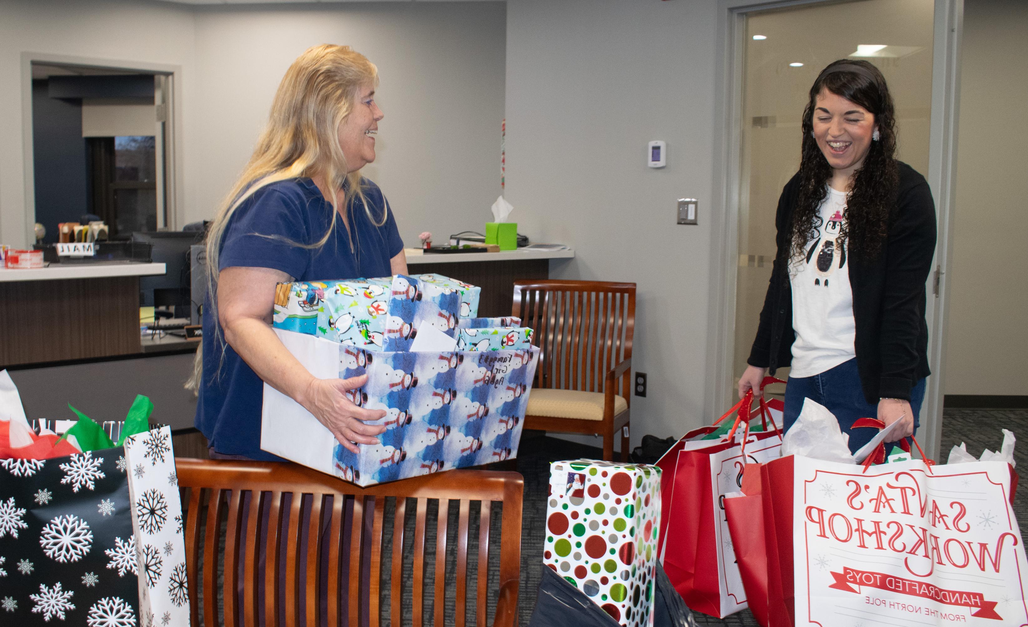 A female employee with long dark hair is smiling and holding several colorful bags filled with gift. Another female employee with long light colored hair is smiling and holding a box decorated with wrapping paper that is filled with gifts.